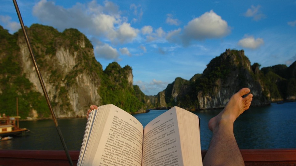 Bare feet and an open book on a boat, with the limestone islands of Halong Bay behind
